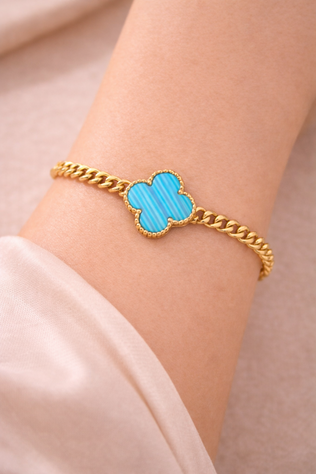 Gold bracelet with a blue clover-shaped clasp on a wrist against a neutral background